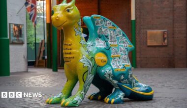 A large dragon sculpture that is green, yellow and blue, with yellow claws. It has drawn stickers of people on it, with a yellow circular logo that reads "Compton Care".