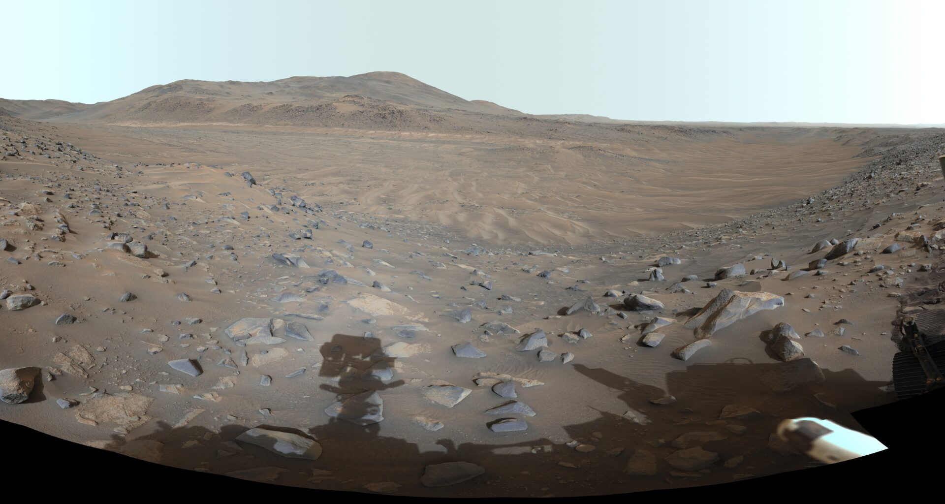 A panoramic view of a barren, rocky landscape with distant hills, framed by parts of a robotic rover visible on the left and right edges.