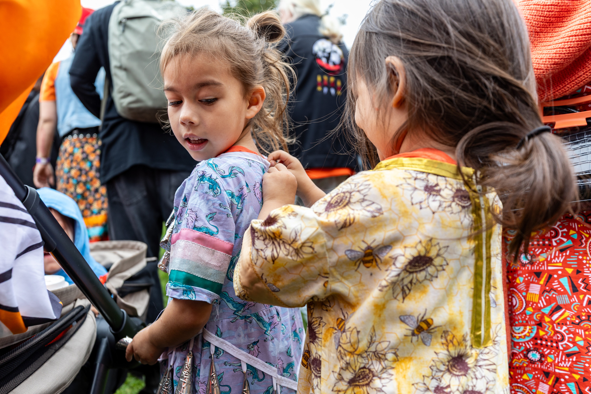 A girl zips up the dress of another at an outdoor gathering.