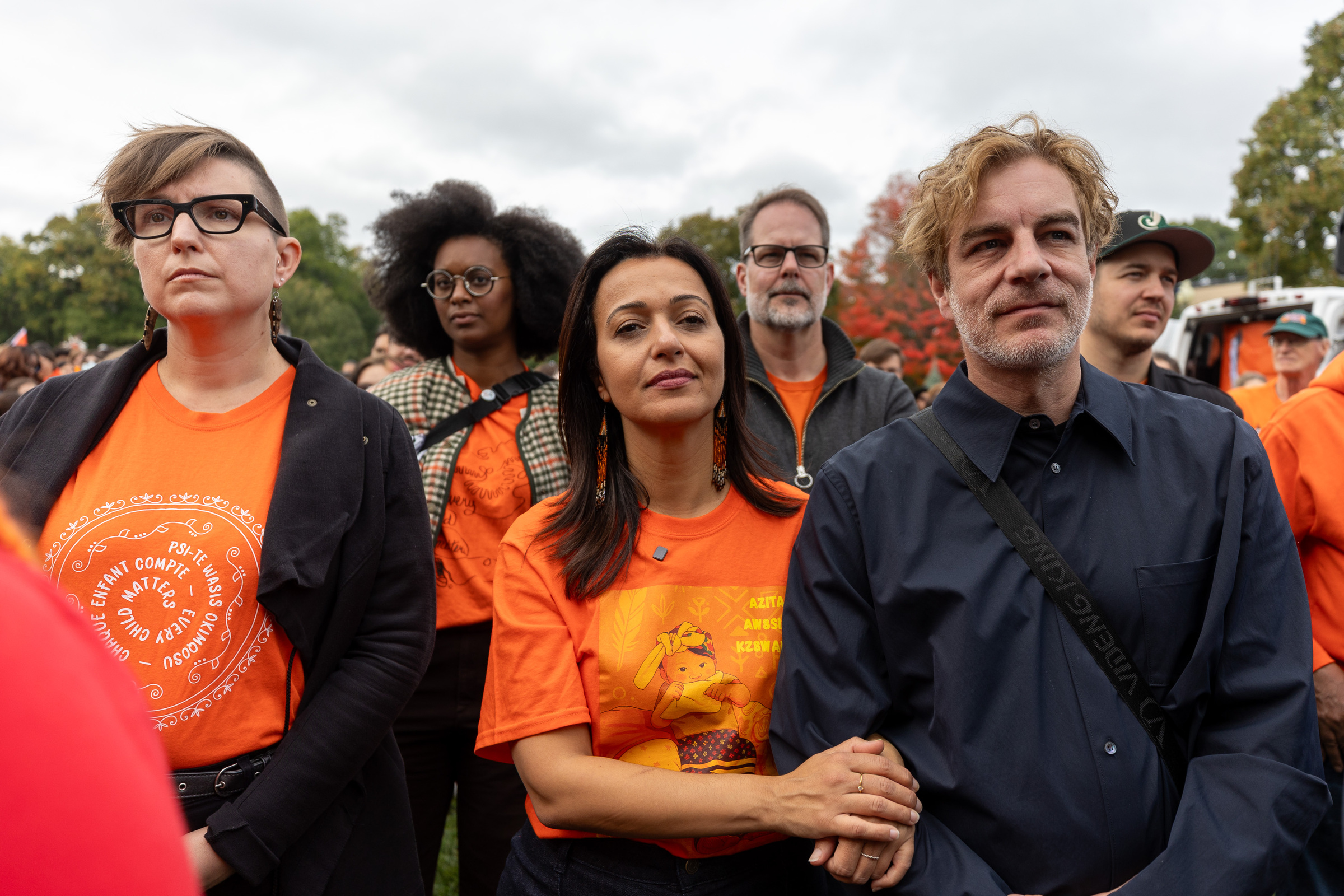 People in orange T-shirts stand and watch an event outdoors.