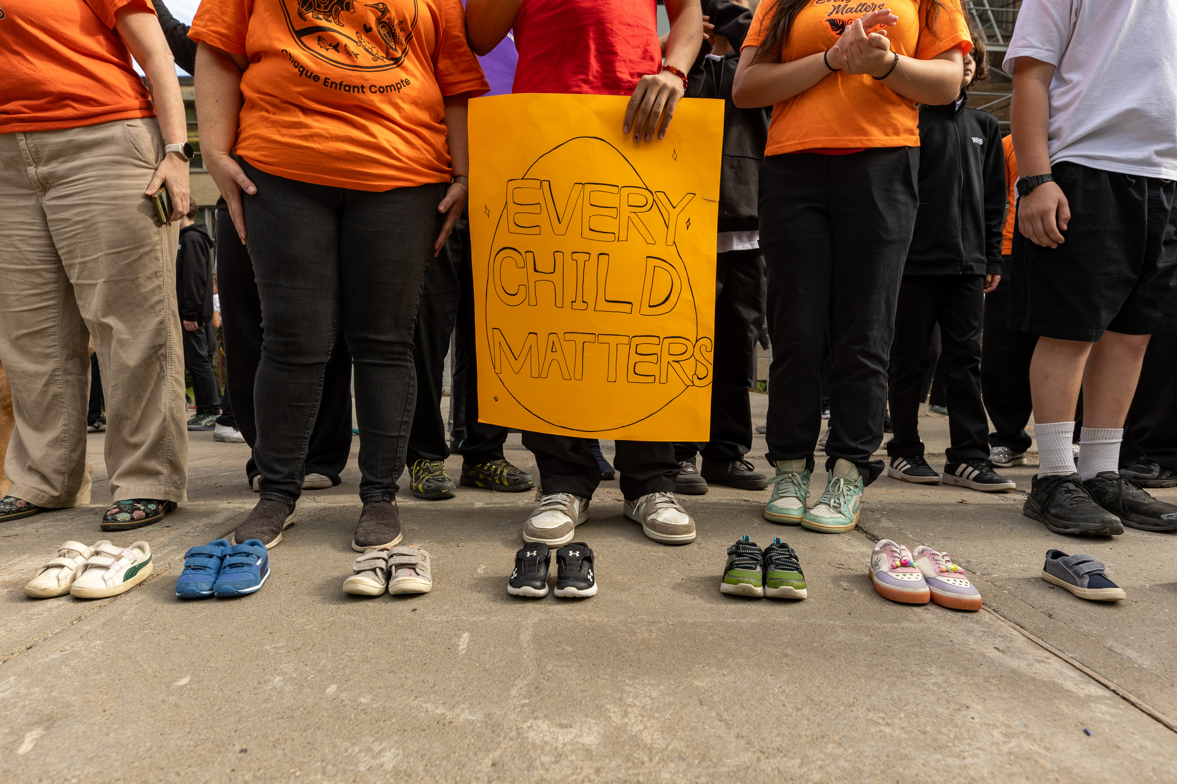 Pairs of shoes are seen lined up neatly on the ground in front of a group of people, one carrying a poster reading Every Child Matters.