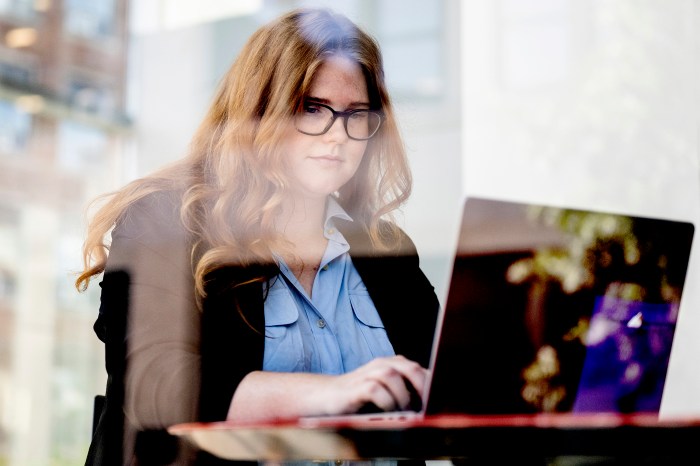 Savannah Reid works on a laptop at a table, seen through a glass window. 