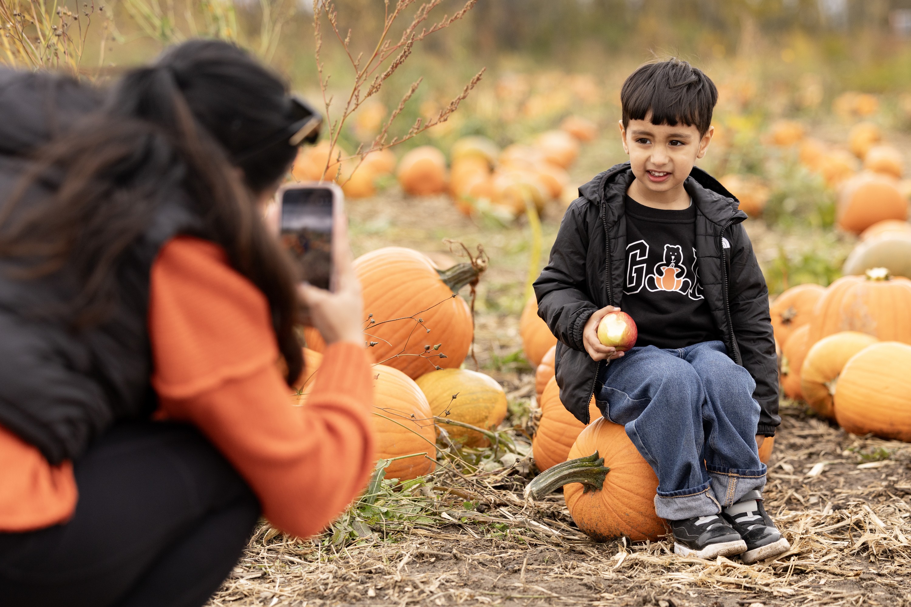 A boy sits on a pumpkin, holding an apple with one bite out of it, while his mother takes a photo using her phone.