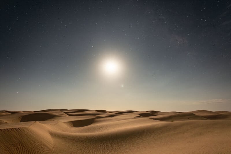A bright moon illuminates a vast desert landscape with rolling sand dunes under a starry night sky.