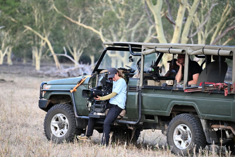 A person sits on the side of a green safari vehicle, operating a professional video camera, while another person sits in the back seat.