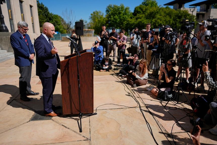two men in suits stand at a podium in front of a crowd of media