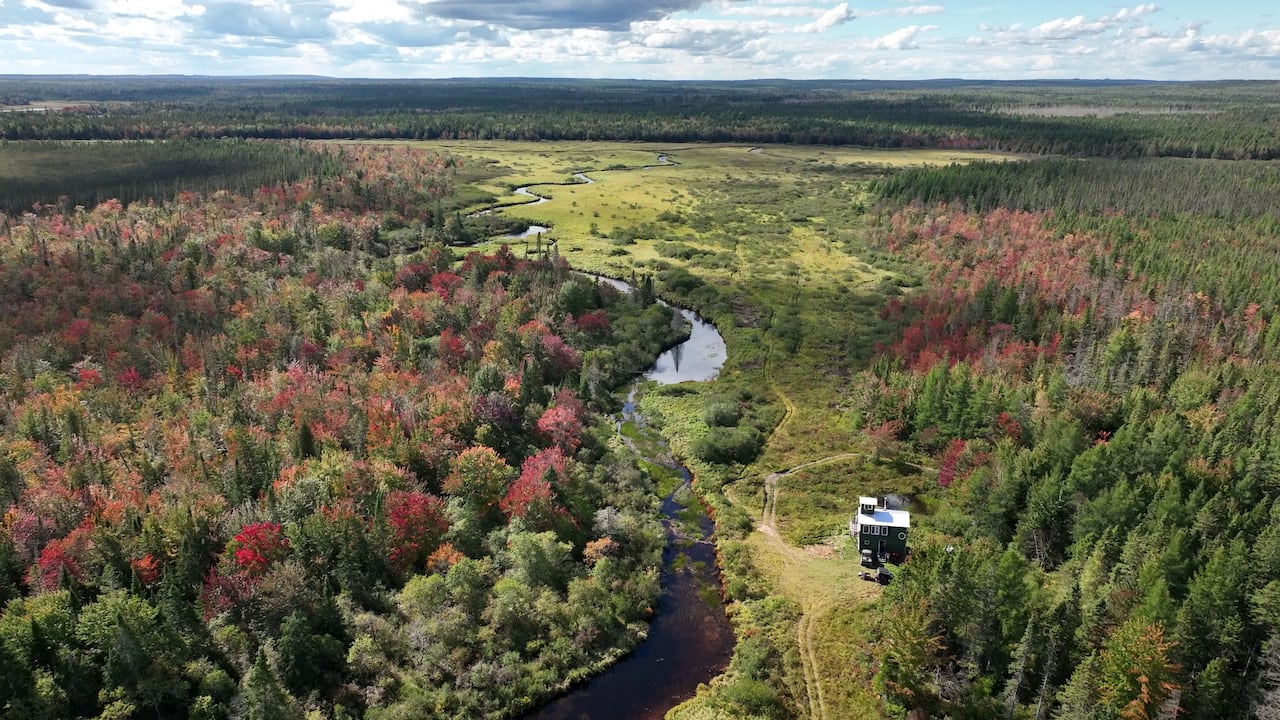 A drone shot of a two storey hunting cabin with a crows nest on top. It sits alongside a stream overlooking meadows among bright fall foliage.