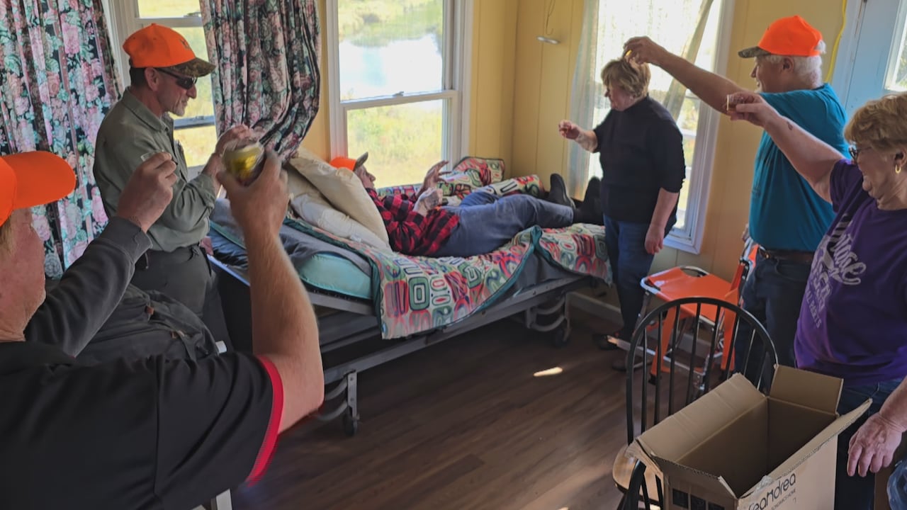 A group of hunters raise a glass in a hunting cabin while one older man wearing a red and black plaid jacket rests in a bed.