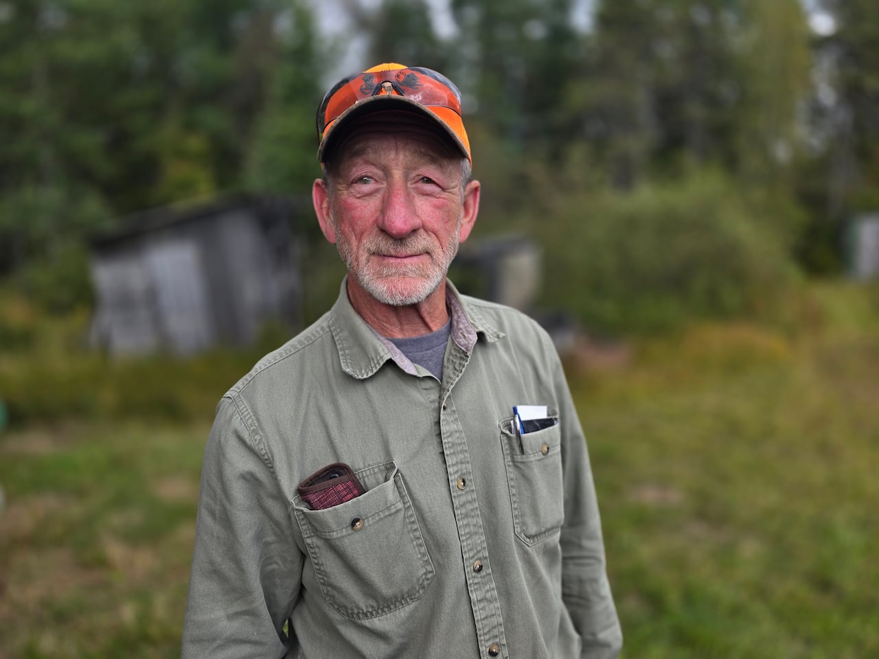 An older white man with a short grey beard standing in the woods wearing a green shirt and an orange hunting cap.