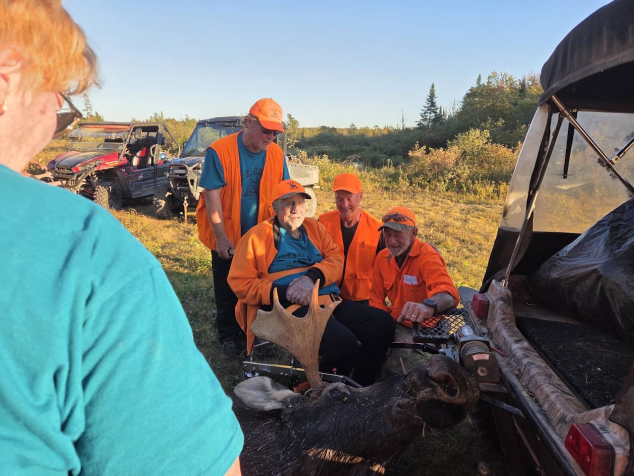 A group of four hunters all wearing orange hunting gear sit around a dead moose hitched to rear of an Argo all-terrain vehicle.