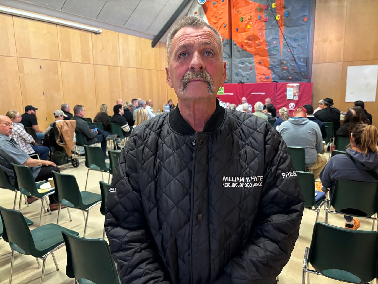 Darrell Warren is wearing a black jacket.  He's standing in a room with people attending the meeting sitting behind him.