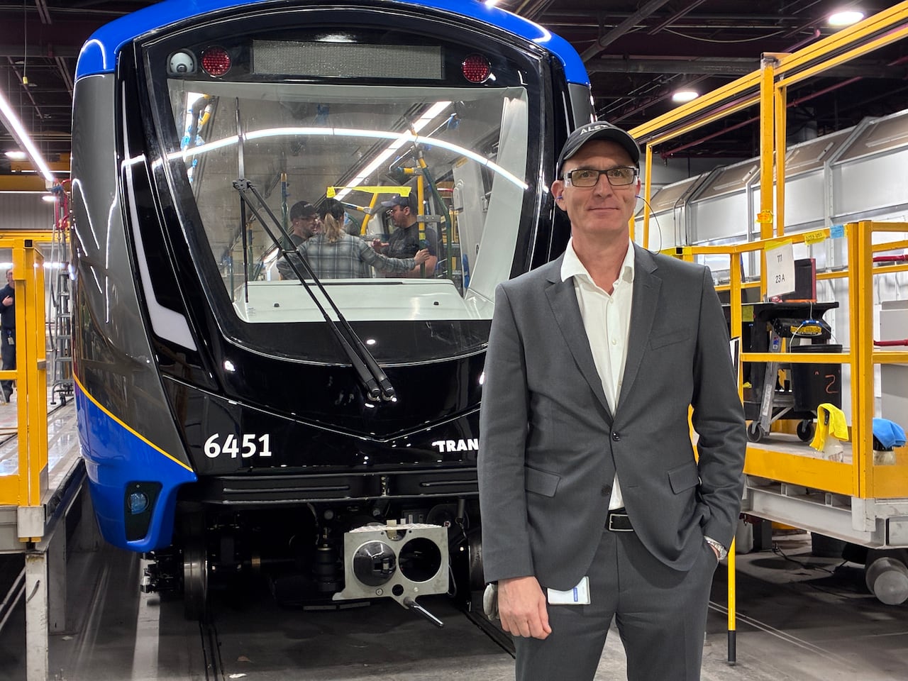 A man in a suit and glasses stands in front of a train car under construction.