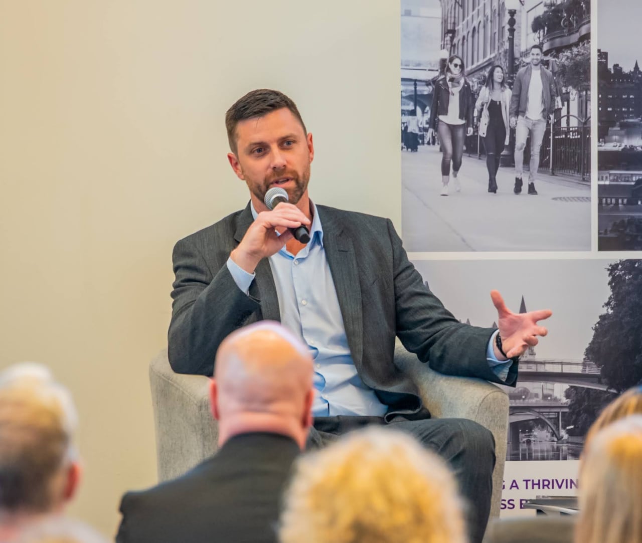 A mayoral candidate speaks at a debate. He's seated in a chair and gesturing with his hand that isn't holding a microphone.