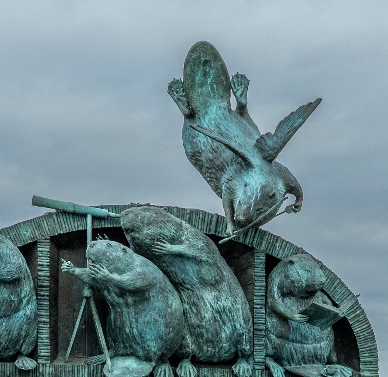 Against a gray sky, a bronze sculpture shows a number of beavers inhabiting a structure. Two look out a telescope while another reads a book. One beaver with wings and an arrow, like cupid, flies overhead.