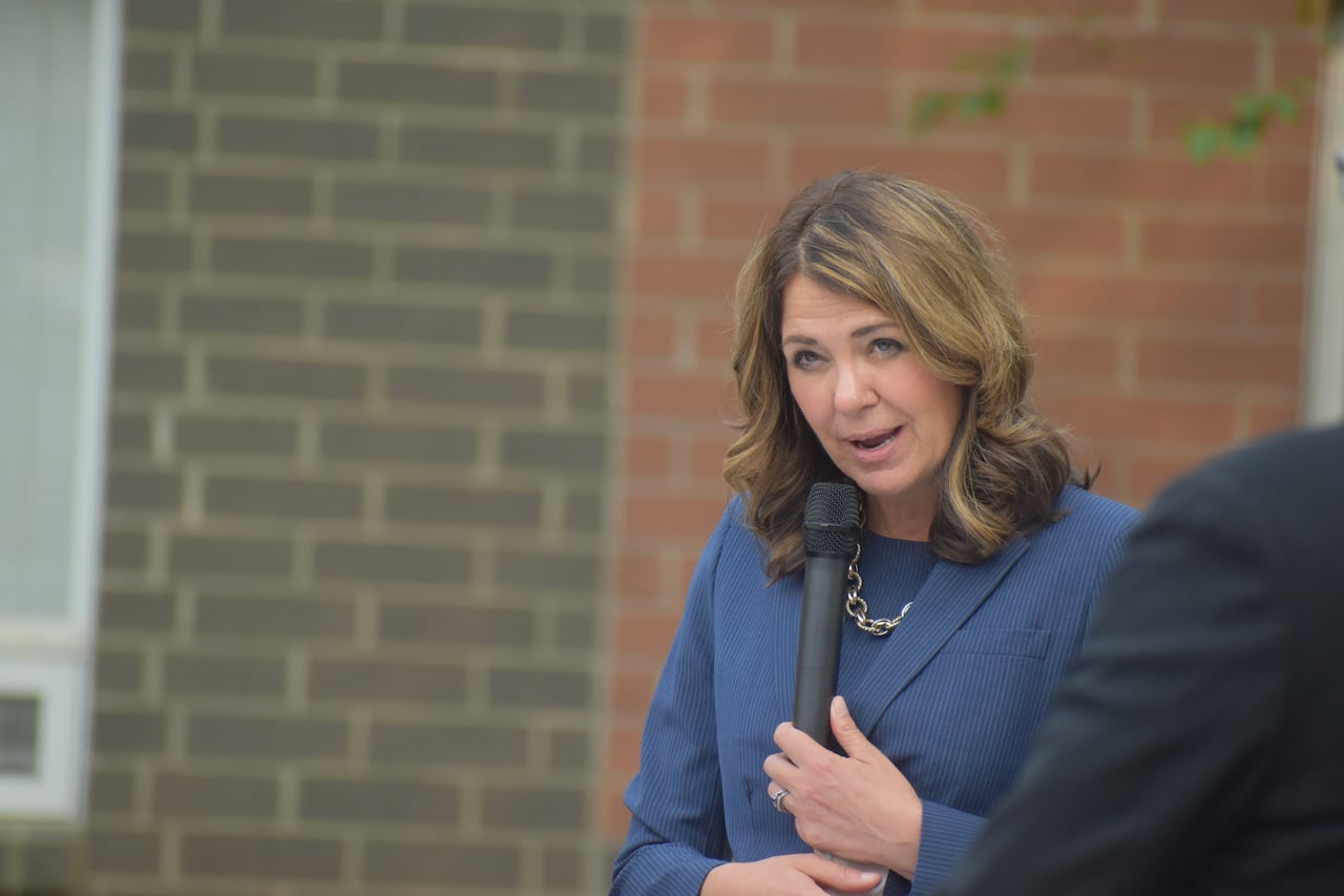 A woman speaks into a microphone in front of a dark, grey brick wall.