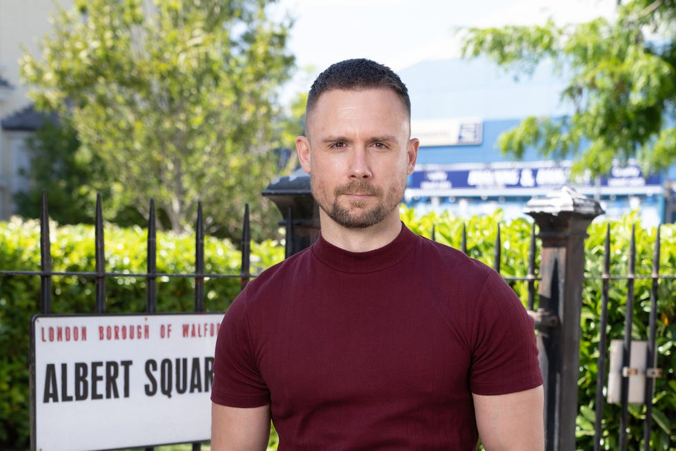 EastEnders, Lee Carter in 2026 eastenders, danny hatchard as lee carter in 2026, wearing a red tshirt standing outside the square gardens