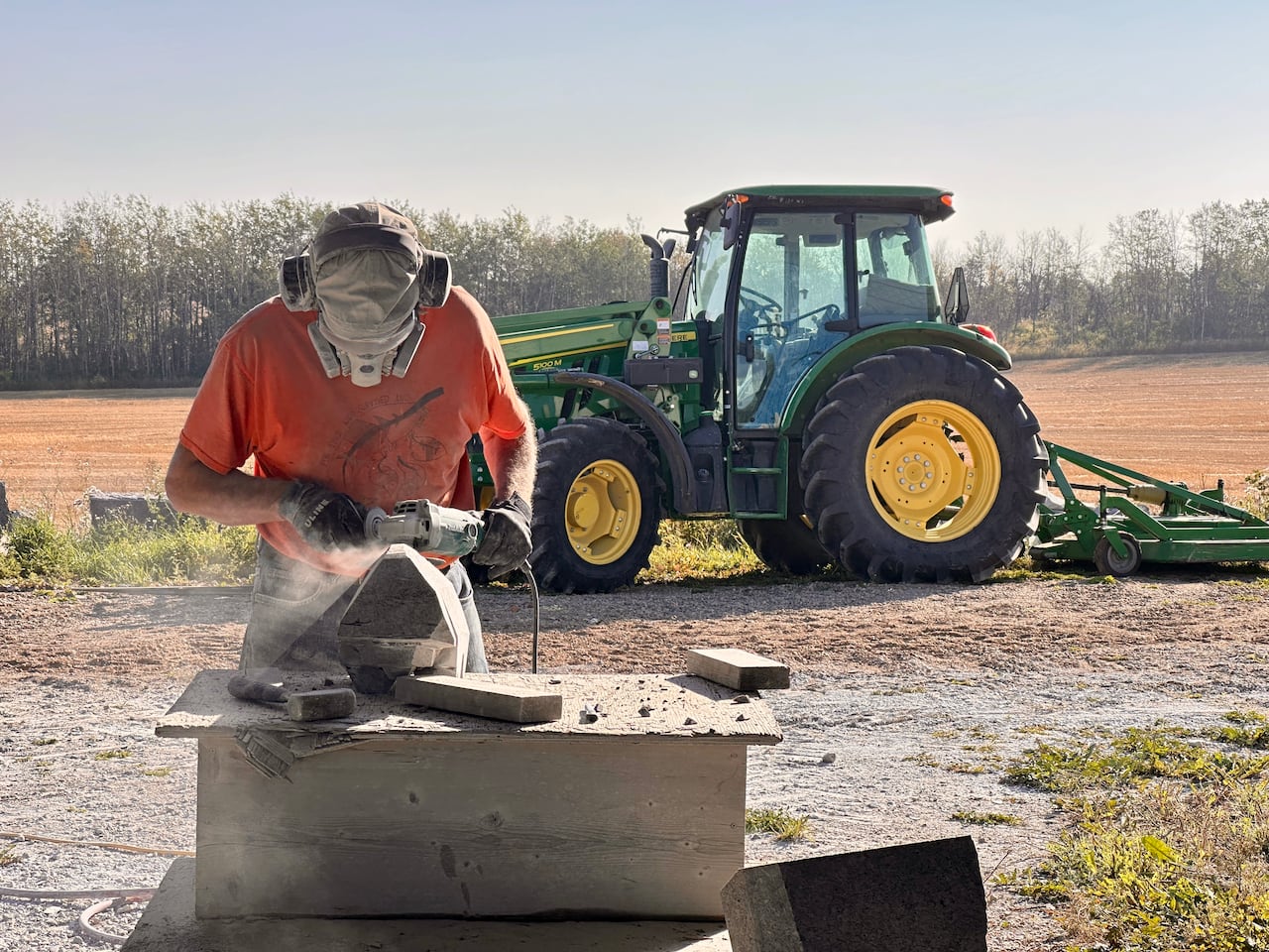 A man in an orange shirt and a face mask works with a stone saw on a piece of rock with a green tractor in the background.