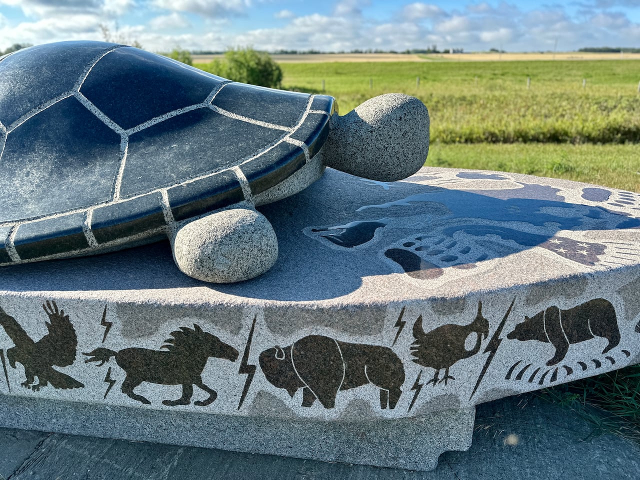 A large carving of a turtle sits on a another stone covered in carvings of horses, bison and bears, set against green grass and a blue sky.