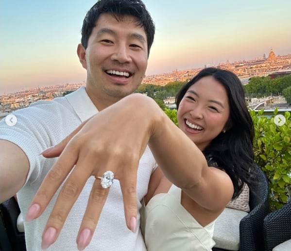 A smiling man and woman pose for a selfie photograph, as she holds up her left hand to the camera to show off a large diamond engagement ring.