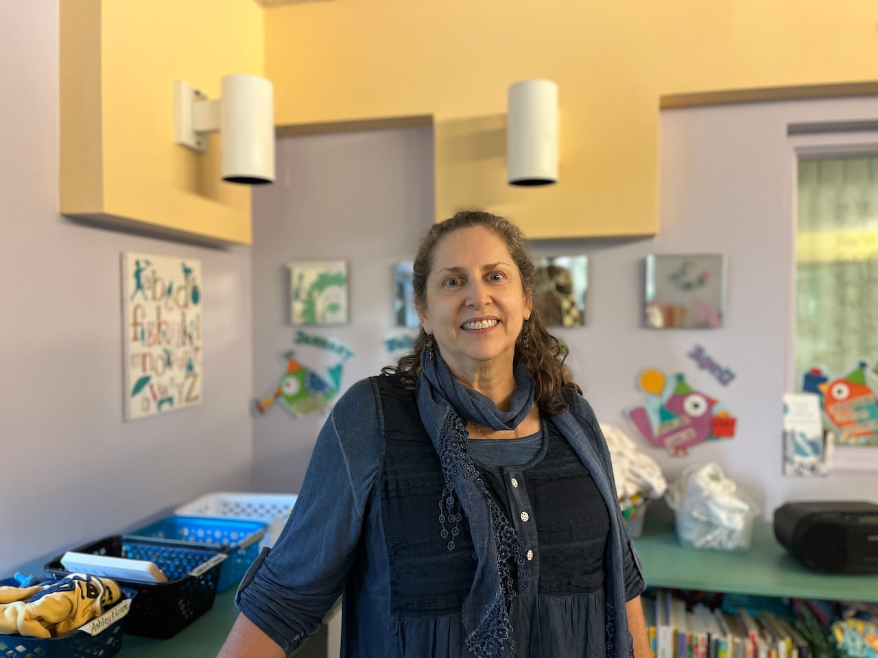 A woman with long grey hair and a blue blouse leans against green shelves filled with children's books.