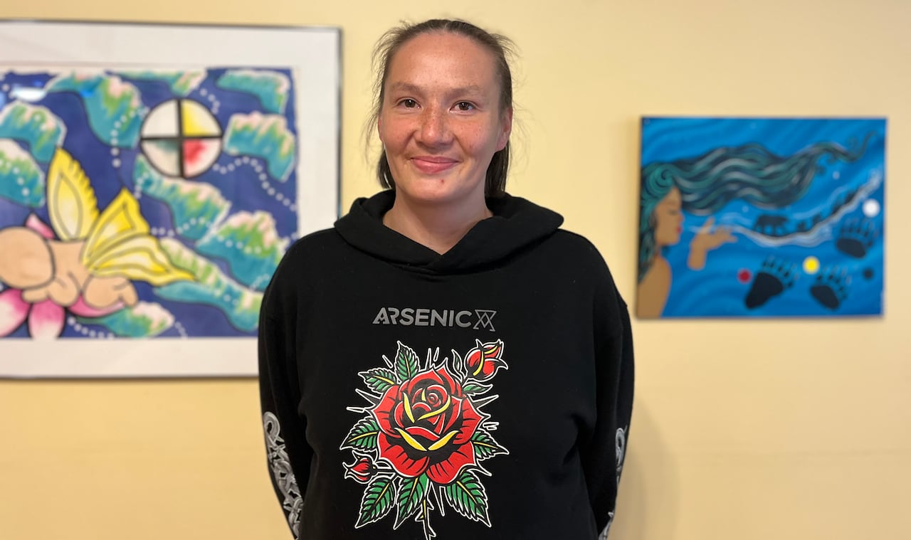 A woman with short hair and a black sweater stands in front of a yellow wall bearing artwork.