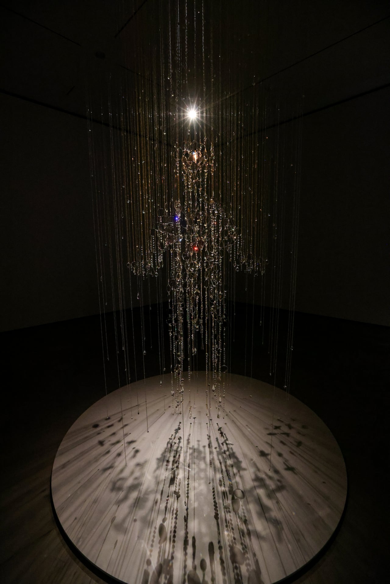 An art installation made of glass and crystal on strings hangs illuminated by a spotlight, creating glimmering reflections and shadows. The glass beads suggest a spectral human form suspended in the air.