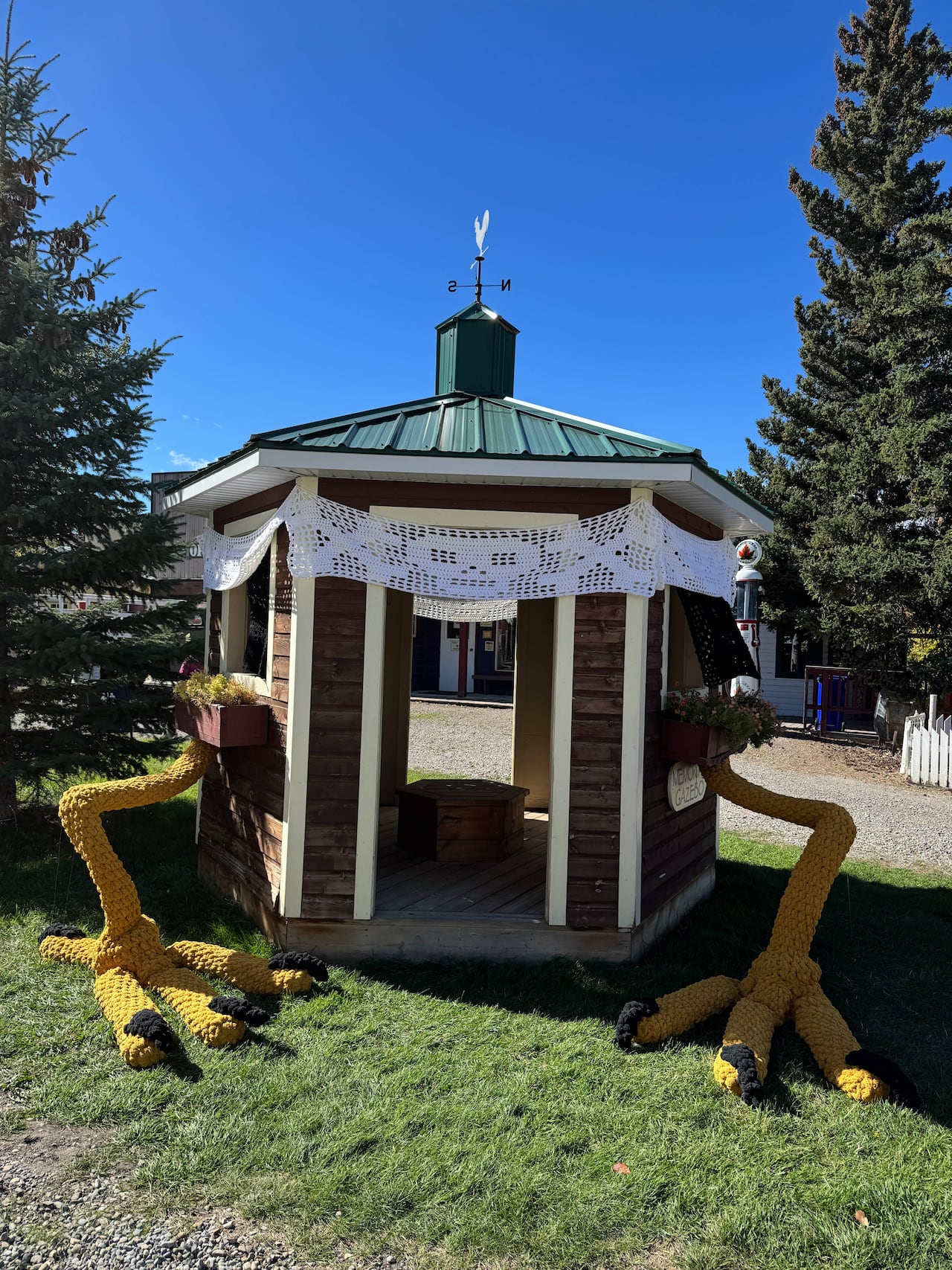 Daytime photo. A shed with a green roof and weather vane has been decorated with textile art to suggest it is a Baba Yaga hut with long yellow bird legs.