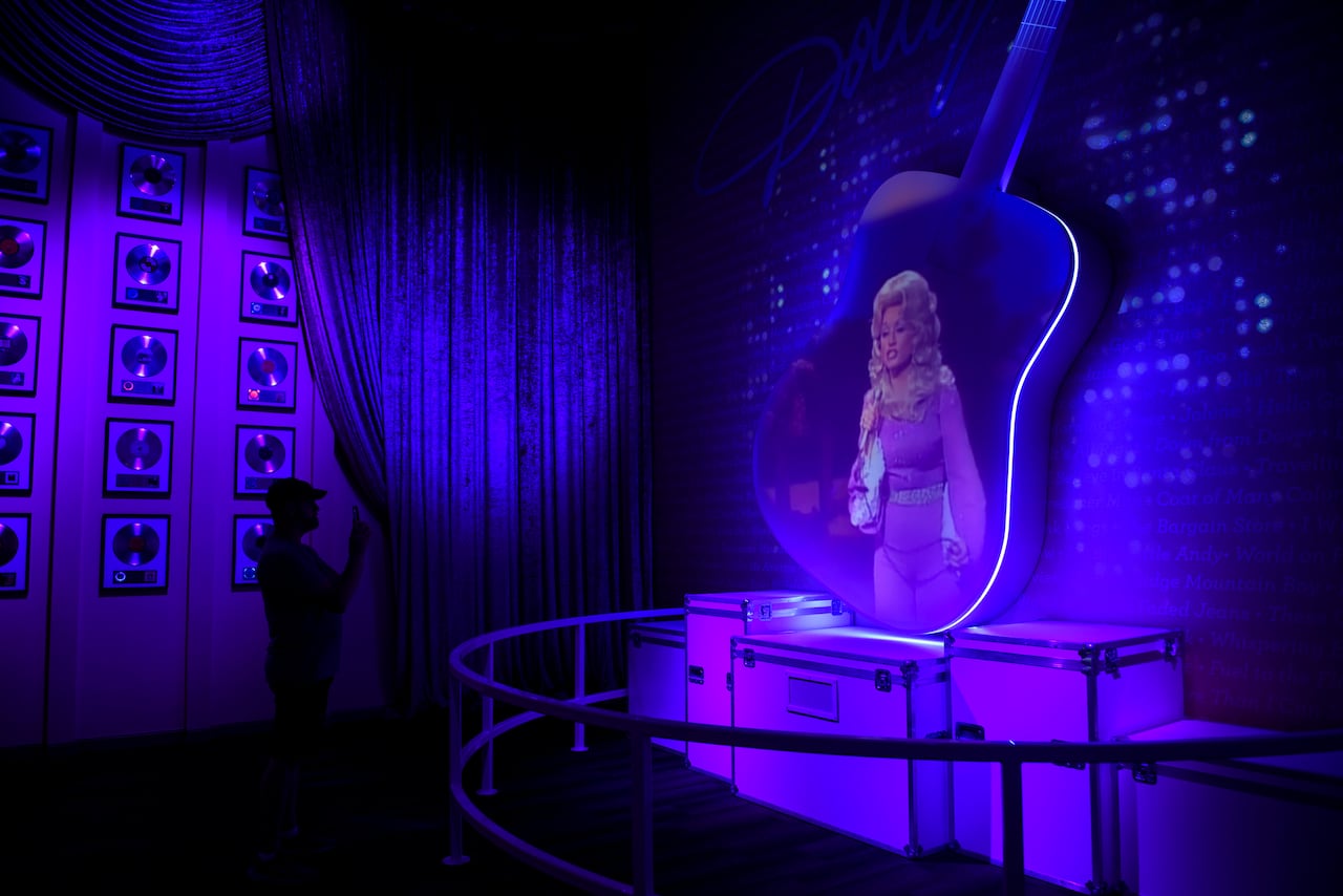 The silhouette of a person standing against a wall of golden records can be seen taking a photo of a large guitar with a picture of a woman on it.