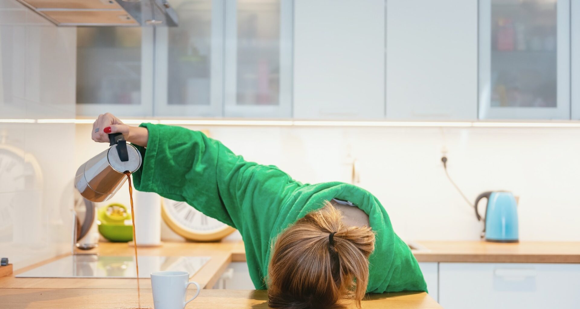Tired woman sleeping on the table in the kitchen at breakfast. Trying to pour morning coffee into mug but spilling on to the counter
