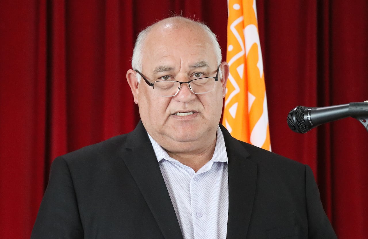 A man in a dark suit jacket with a light blue shirt looks over his glasses while speaking into a microphone.