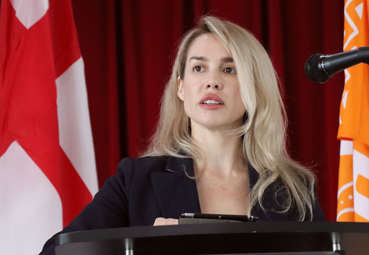 A woman with long blonde hair wearing a dark blazer speaks into a microphone.