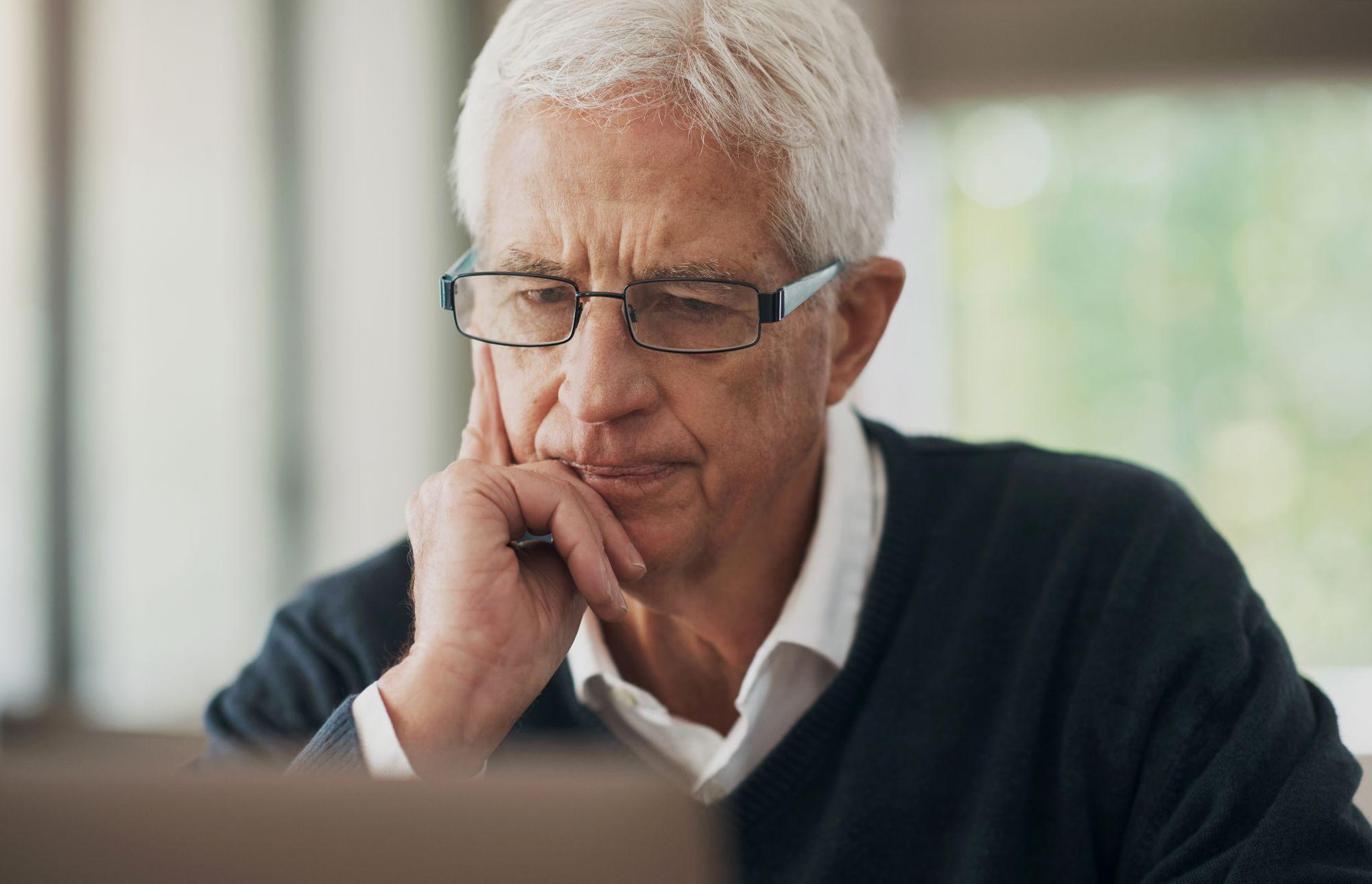 Thoughtful senior man wearing glasses rests his face on his hand while looking at a laptop.