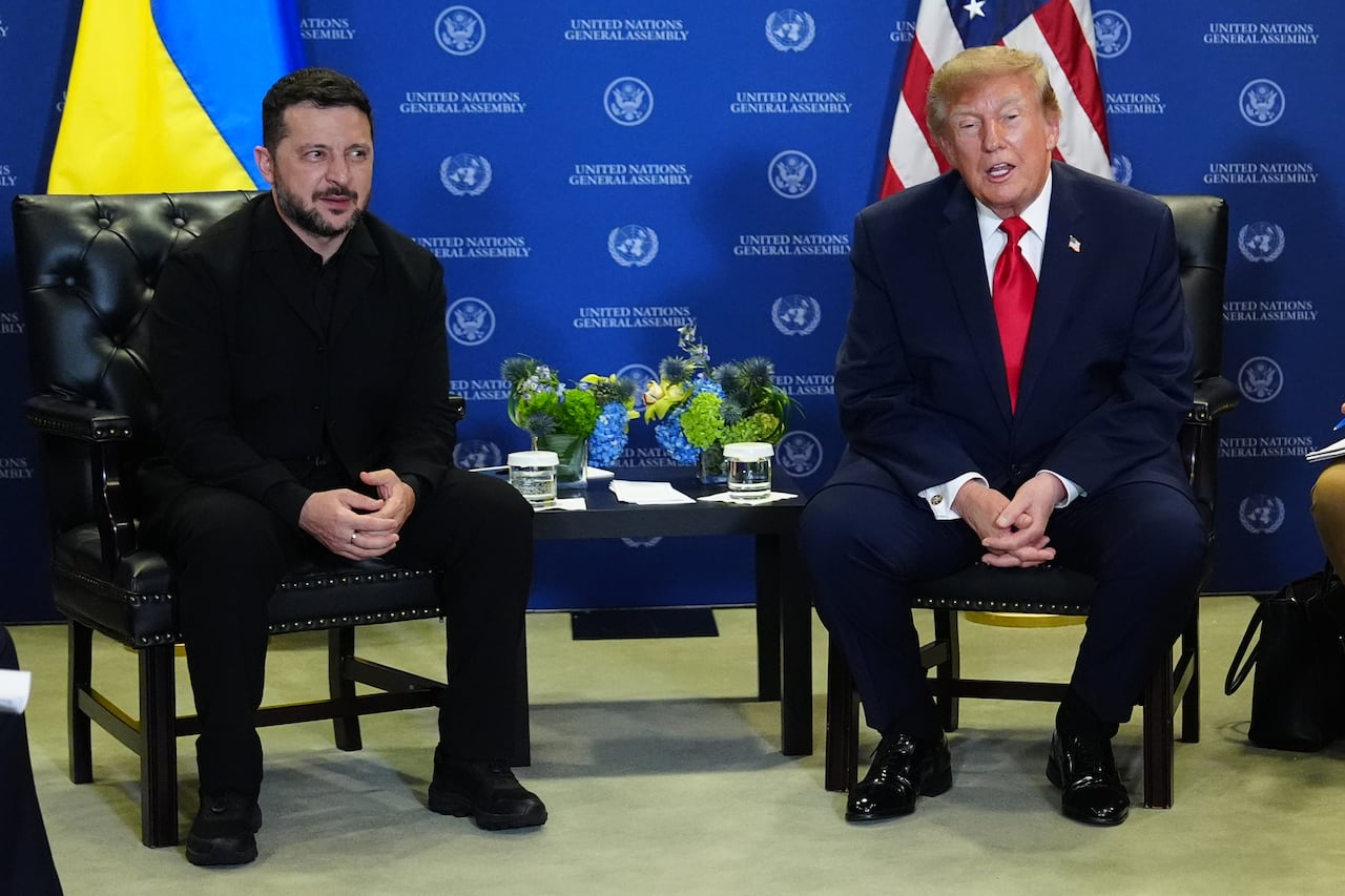 Two men sit on chairs in front of a Ukrainian flag and an American flag.