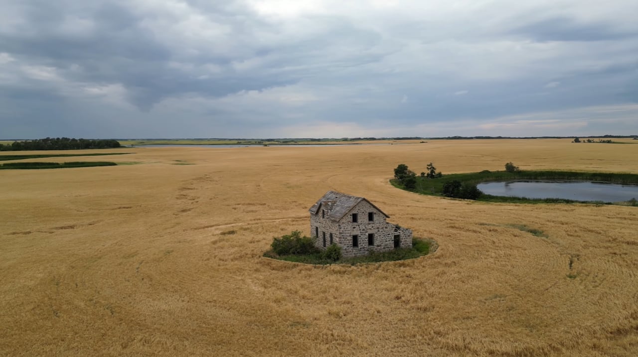 Aerial view of an old stone house in the middle of a ripe wheat field, under a cloudy sky.