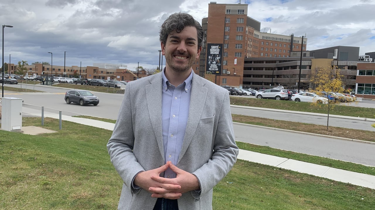 A man in a grey blazer stands outside a hospital on a cloudy day.