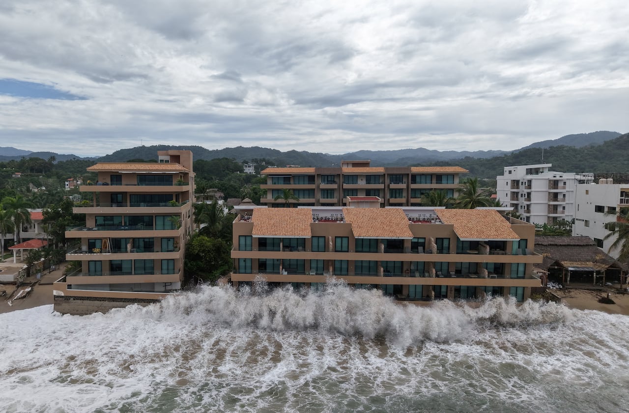 Large waves hit buildings on a coast.