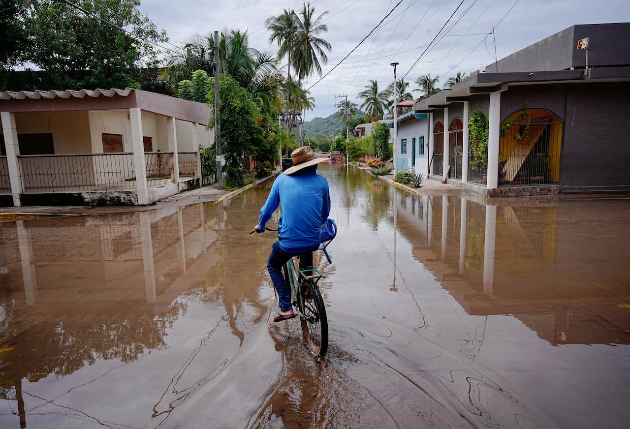 A person in a hat with their back to the camera rides a bike down a road filled with higher-than-normal water on the road.