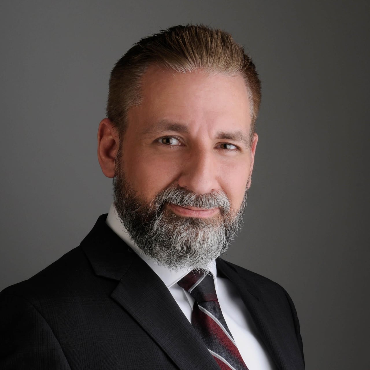 A professional headshot of a man with a beard, wearing a black suit jacket, white button-up shirt and a red, black, silver and white striped tie.