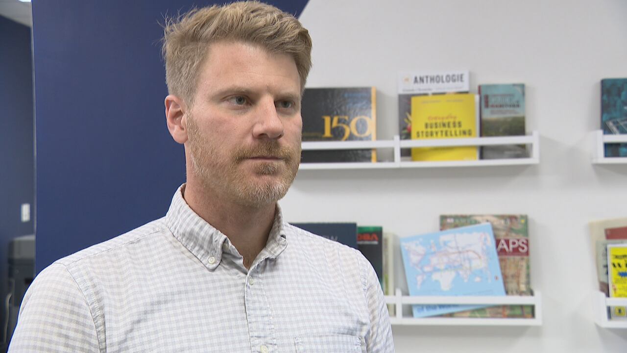A man with short blonde hair parted to one side, and a blonde beard. He wears a collared shirt and stands inside an office.