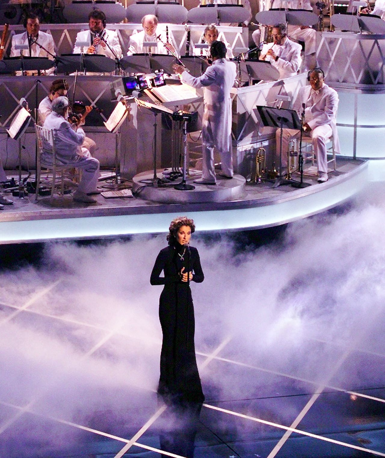 An image captured from above shows a dark-gowned woman singing amid fog onstage, with a conductor-led orchestra dressed in white playing behind her.