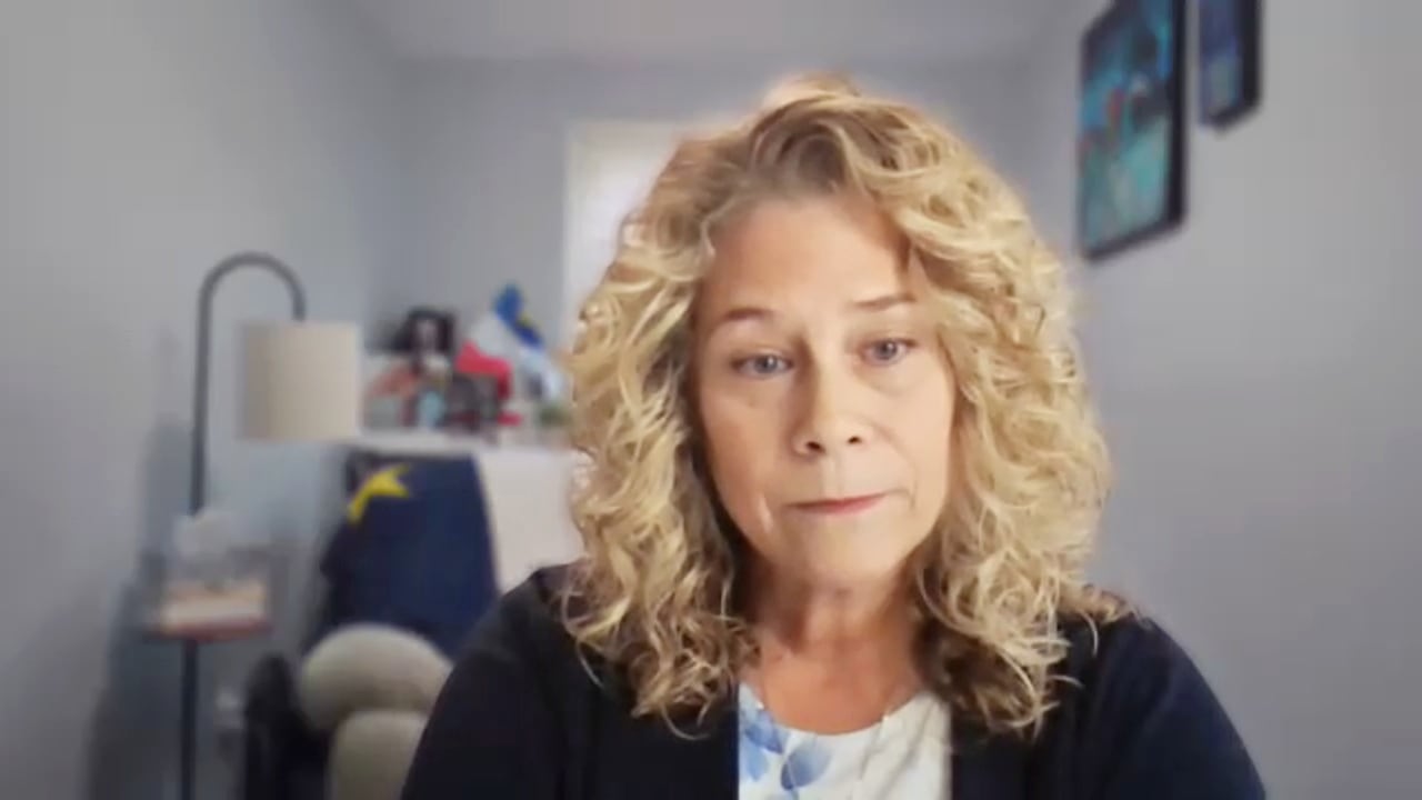 A white woman with curly blonde hair wears a black cardigan, and sits in a room that is slightly blurred behind her.