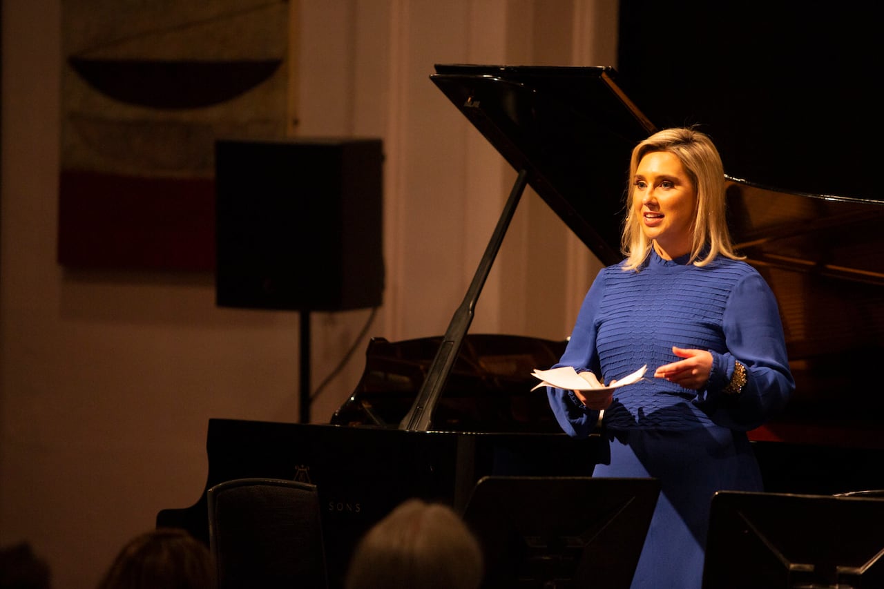 A woman speaks to an audience with a grand piano in the background.