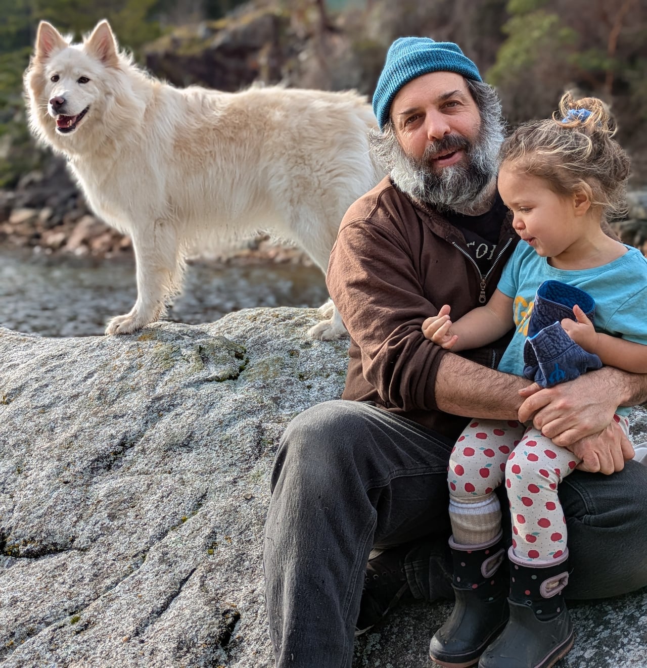 A man with a beard sits on a shoreline with his daughter and dog.