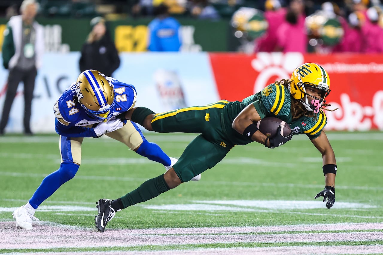 A football player in a green and yellow jersey holds a football as another player in a blue and gold uniform trips him by grabbing on to his foot.