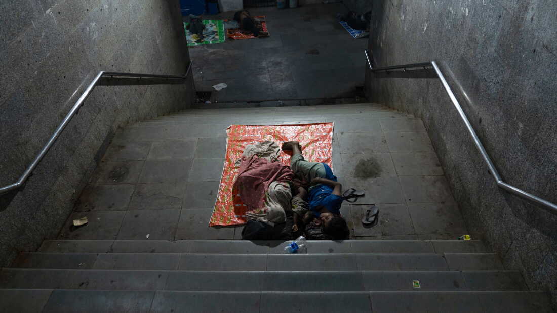 A family sleeps on pavement on a landing in between stairs inside a subway. 