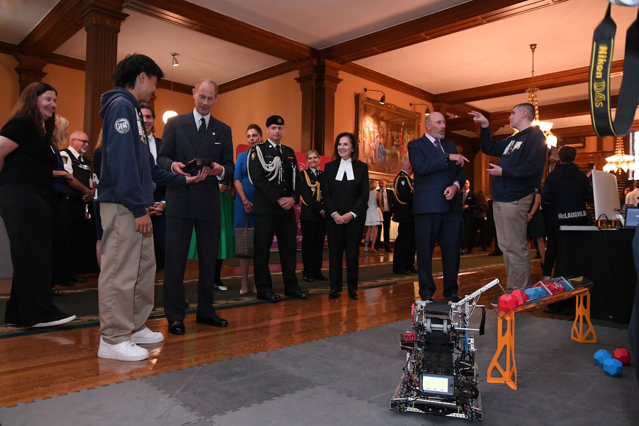 Adults and a teen watch as a robot is deploys an arm to pick up a block.