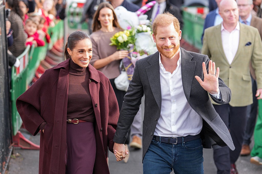 Two people holding hands walk alongside people behind barricades while a person carrying a bouquet of flowers follows.