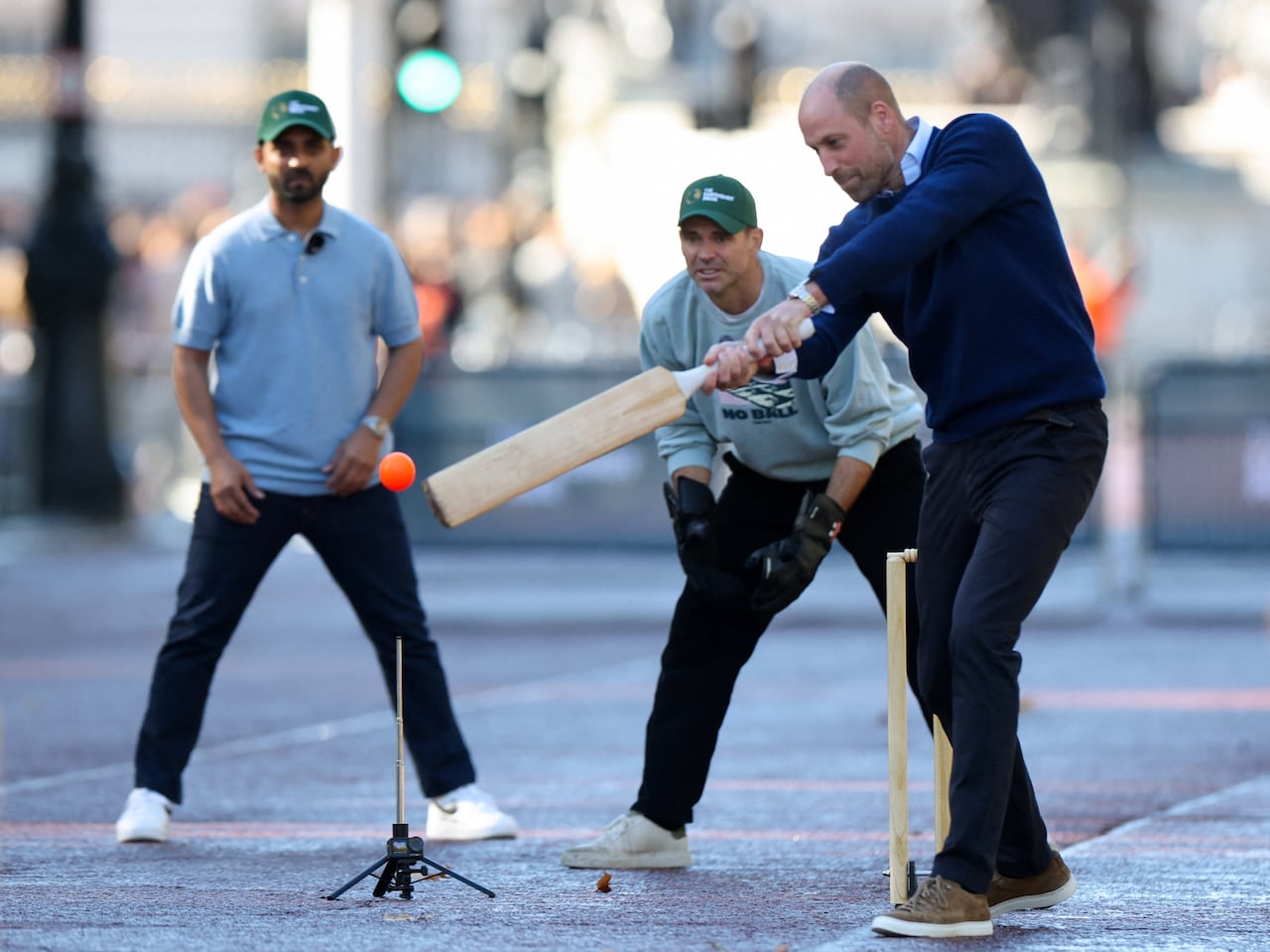 A person swings a cricket bat at a ball as two other people look on.