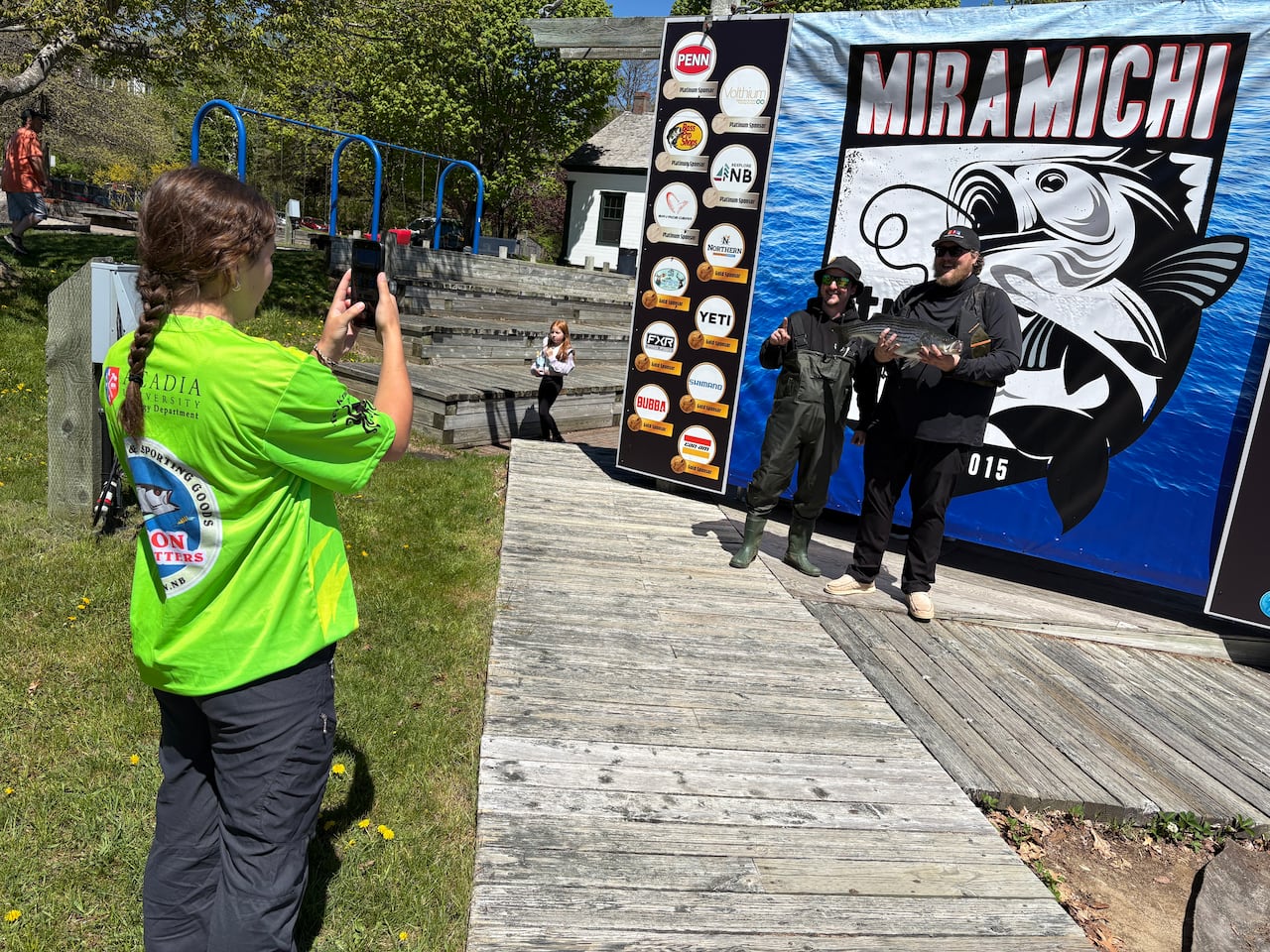 A person takes a photo of two people holding a striped bass, against a backdrop of a poster depicting the fish.