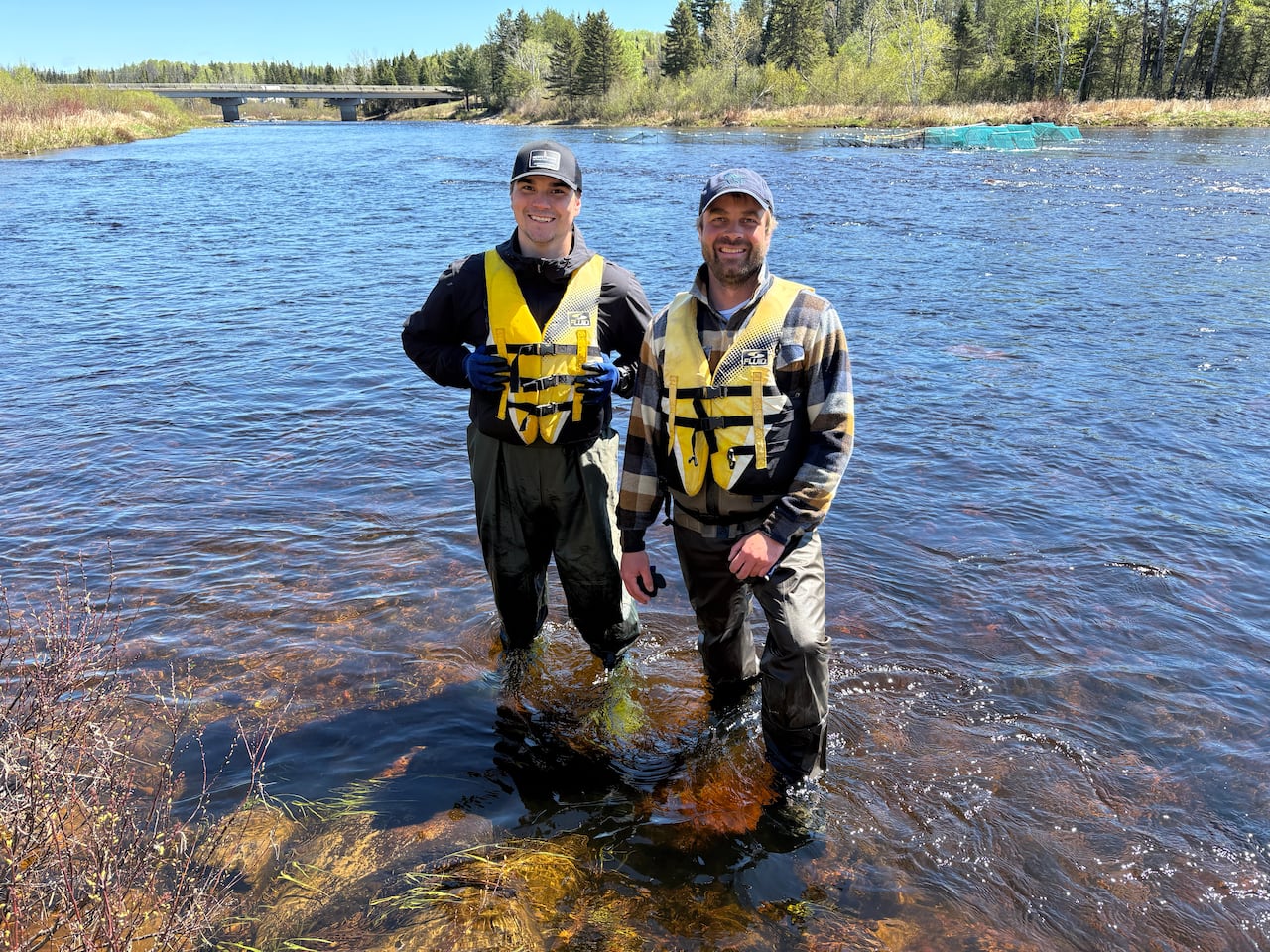 Two men in waterproof fishing gear stand knee deep in a river and smile for the camera.